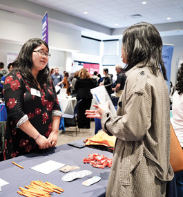 A young woman stands at a table in a conference space, talking with another young woman